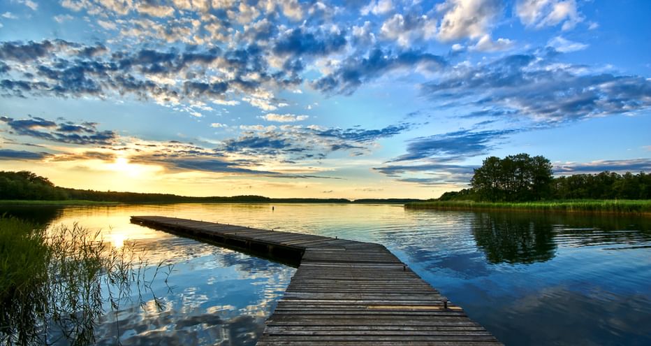 Holzsteg, der sich bei Sonnenuntergang in einen ruhigen masurischen See erstreckt. Der Himmel ist voller dramatischer Wolken, die sich im Wasser spiegeln.