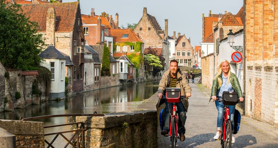 Zwei Radfahrer auf roten Rädern überqueren eine Steinbrücke in einer historischen flämischen Stadt mit Kanal, traditionellen Backsteinhäusern und Ziegeldächern.