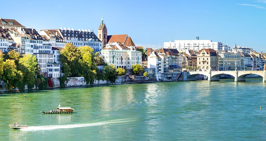 Grüner Rhein mit Booten, historischen Basler Gebäuden, Kirchturm und Steinbrücke unter blauem Himmel. Bäume säumen das Ufer.