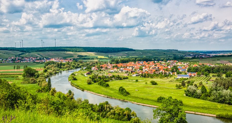 Luftaufnahme eines sich schlängelnden Flusses durch grüne Wiesen mit einem Dorf mit roten Dächern. Hügel und Windräder in der Ferne unter bewölktem Himmel.
