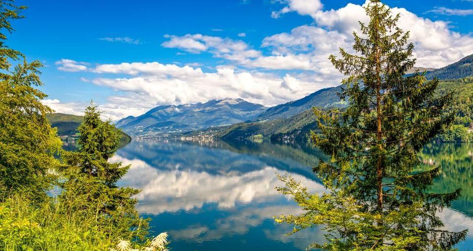 Millstätter See in Austria with calm blue water reflecting mountains and white clouds. Green trees frame the view on both sides under a bright blue sky.