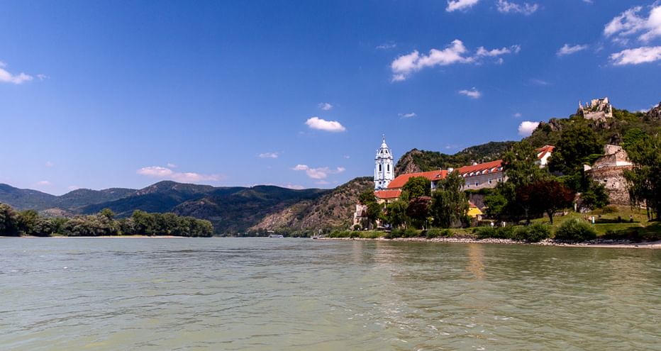 View of the Danube River with Dürnstein Abbey's blue-white tower and castle ruins on a hilltop. Mountains and blue sky in the background.