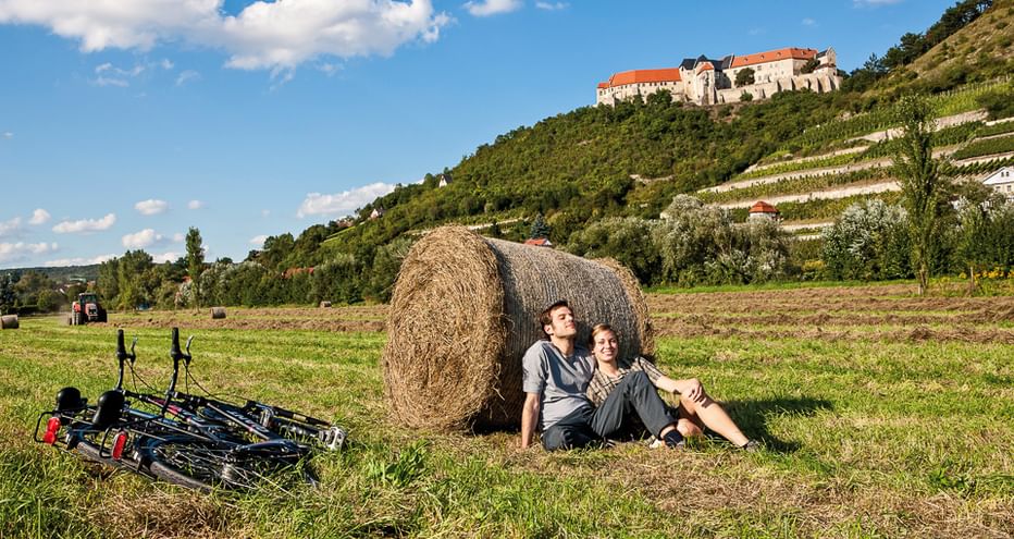 Zwei Radfahrer lehnen an einem Heuballen auf einer Wiese, Fahrräder daneben. Weinberge und eine Burg auf dem Hügel im Hintergrund.