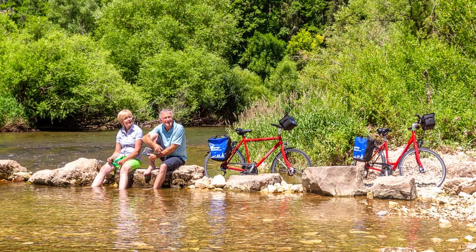 Two cyclists sitting on rocks by the Danube River near Fridingen with their red touring bikes. Limestone cliffs and green forest in background.