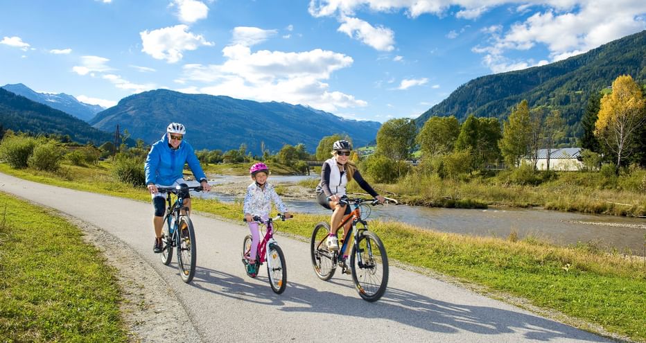 Familie mit drei Personen radelt auf asphaltiertem Weg entlang der Mur mit Bergen im Hintergrund. Erwachsene und Kind tragen Helme an sonnigem Tag.