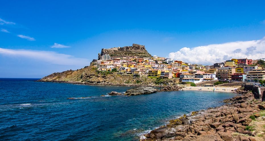 Bunte Hangstadt mit Festungsruinen auf dem Gipfel mit Blick auf eine blaue Mittelmeerbucht mit felsiger Küste in Nordsardinien.