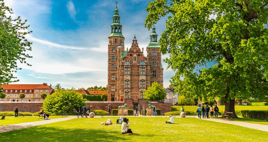 Schloss Rosenborg mit roten Backsteintürmen und grünen Kupferspitzen in Kopenhagen, umgeben von Parkrasen mit Besuchern unter blauem Himmel.