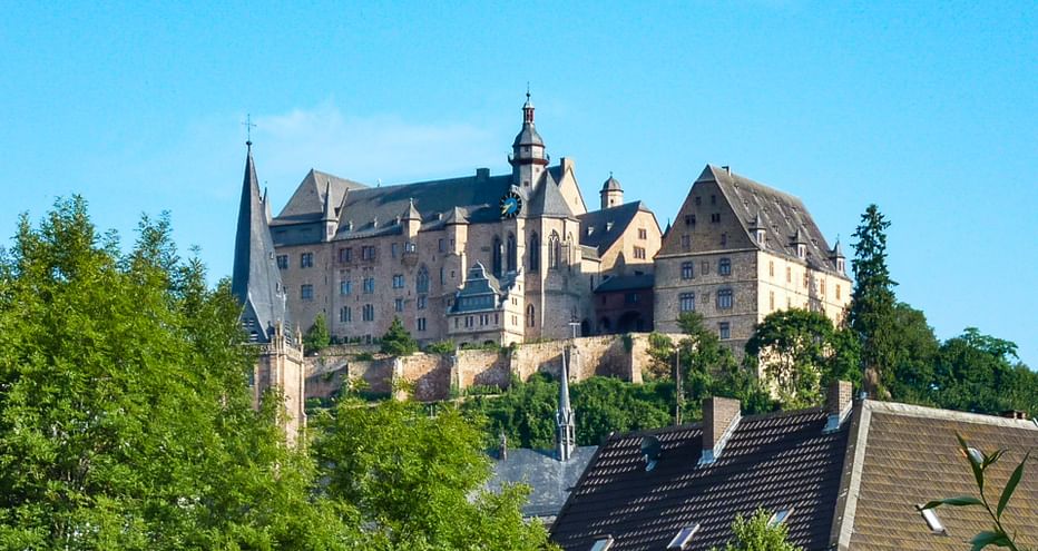 Historic Marburg Castle perched on a hilltop with stone walls and towers, surrounded by green trees. Town rooftops visible below against a clear blue sky.