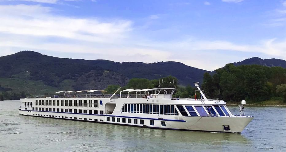 White river cruise ship with blue accents sailing on the Danube. Green hills and mountains visible in the background under a blue sky.