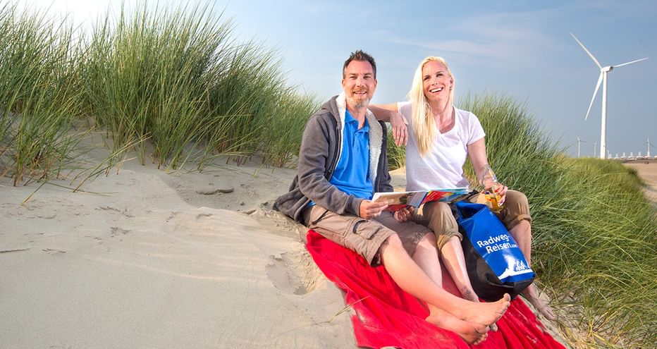 Lächelndes Paar sitzt auf roter Decke in Dünen in Zeeland mit Radkarte. Dünengras und Windräder im Hintergrund sichtbar.