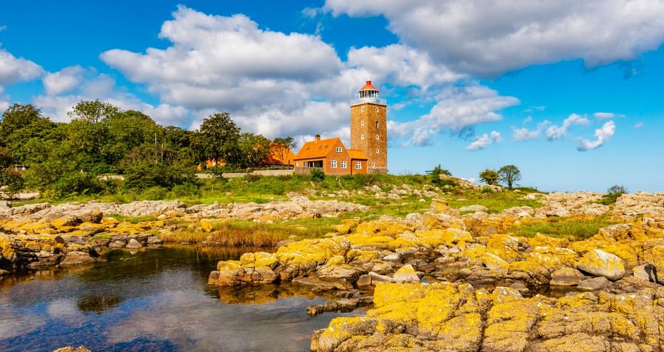 Leuchtturm Svaneke mit rotem Ziegeldach auf felsiger Küste in Bornholm. Gelb bewachsene Felsen im Vordergrund, blauer Himmel mit Wolken.