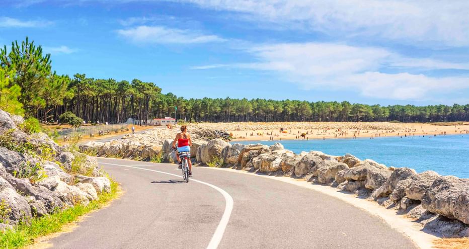 Radfahrerin im roten Shirt auf asphaltiertem Küstenweg bei La Palmyre. Felsige Küste, blaues Wasser, Sandstrand und Pinienwald im Hintergrund.
