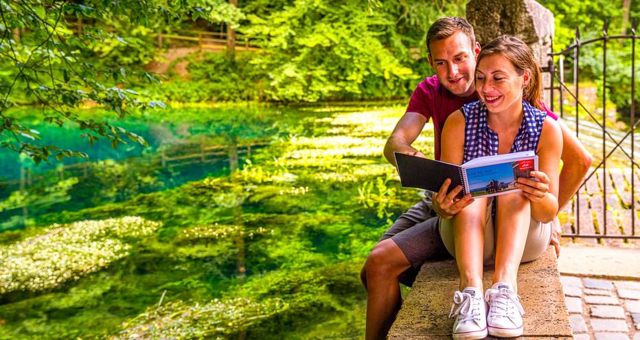 Paar sitzt am Rand des leuchtend grünen Blautopfs in Blaubeuren und liest einen Reiseführer, umgeben von üppiger Vegetation am Wasser.