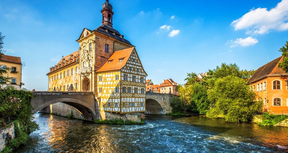 Historic half-timbered Old Town Hall in Bamberg built on a stone bridge over the Regnitz River, with tower and colorful facades under blue sky.