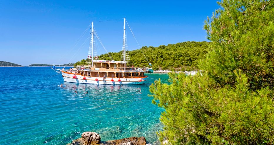 Traditionelles Holzsegelschiff MS Dalmatino vor Anker in kristallklarer türkisfarbener Bucht in Dalmatien, Kroatien, umgeben von Pinien und Felsküste.