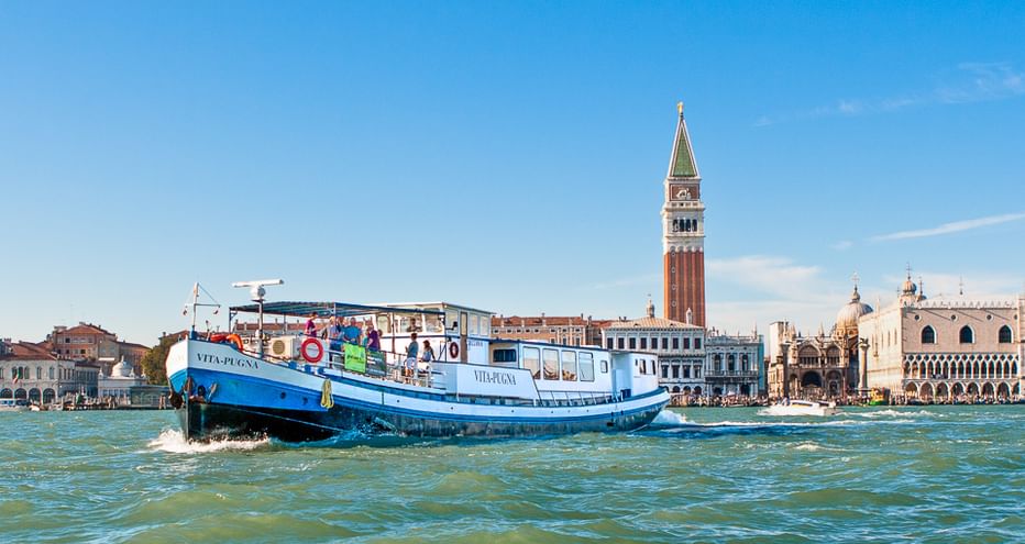 Blue cruise ship sailing on water in Venice with St. Mark's Campanile and historic buildings visible in the background under clear blue sky.