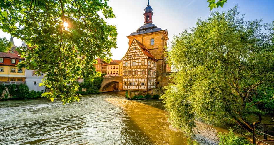 Historisches Fachwerkrathaus von Bamberg auf einer Insel in der Regnitz, umrahmt von grünen Bäumen mit durchscheinendem Sonnenlicht.