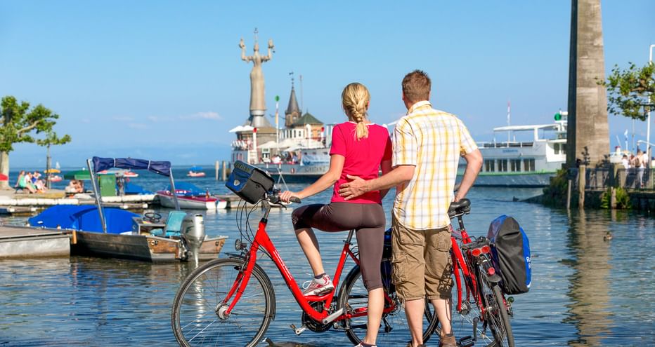 Paar mit Fahrrädern betrachtet den Hafen von Konstanz am Bodensee. Fähre und Imperia-Statue sichtbar, Boote im sonnigen Hafen.