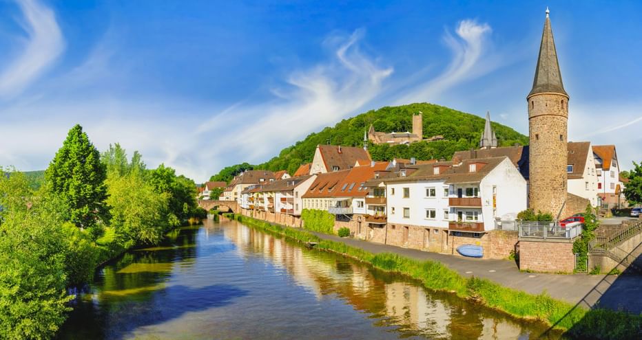Historischer Hexenturm am Fluss in Gemünden mit traditionellen Häusern am Ufer und der Scherenburg im Hintergrund.