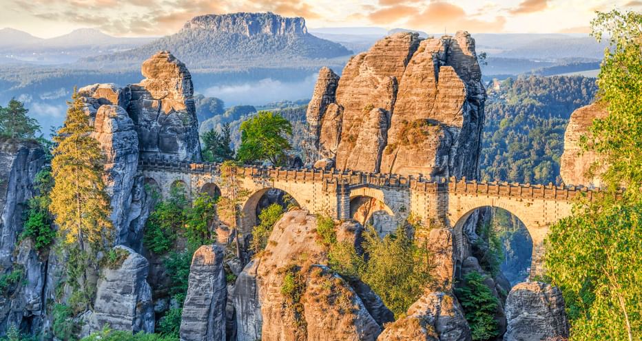 Historische Basteibrücke über dramatischen Sandsteinfelsen im Elbsandsteingebirge bei Sonnenaufgang mit bewaldeten Tälern.