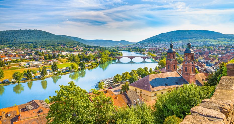 Luftaufnahme von Miltenberg mit rotgedeckten Gebäuden, Kirche mit zwei Türmen, Main mit Steinbrücke und grünen Hügeln unter blauem Himmel.