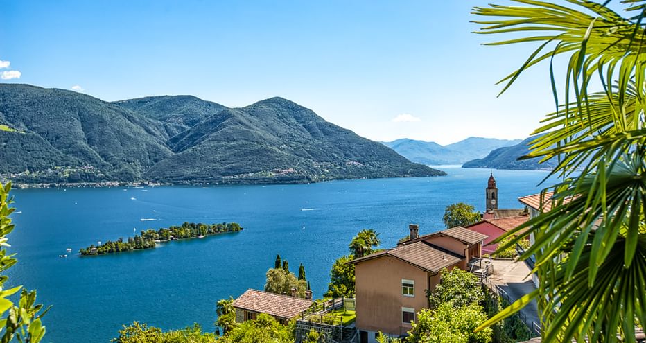 Malerischer Blick auf den Lago Maggiore mit blauem Wasser, kleinen Inseln und Bergen. Häuser mit roten Dächern und Kirchturm im Vordergrund, Palmwedel sichtbar.