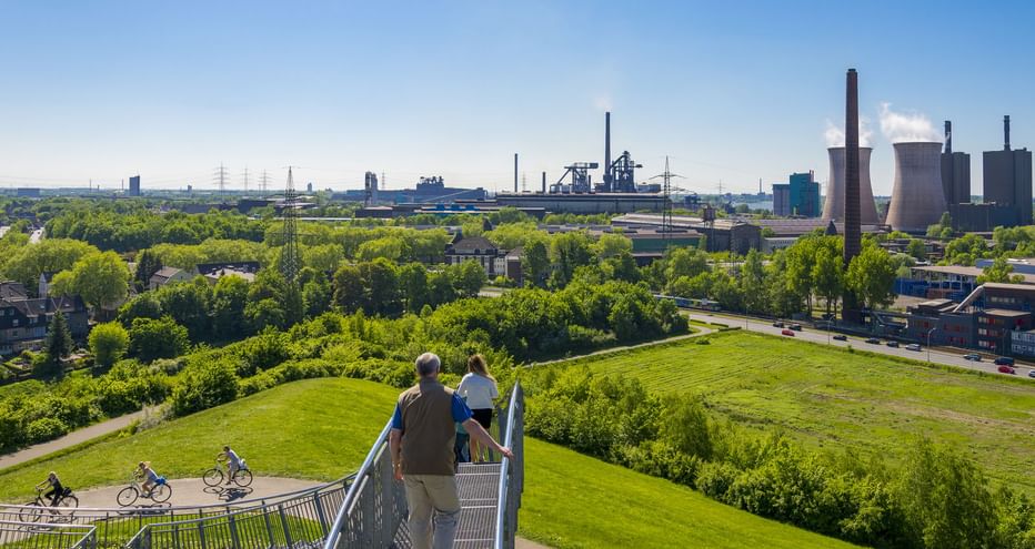 Erhöhter Gehweg im Landschaftspark Duisburg-Nord mit Besuchern, Blick auf grüne Landschaft, Radfahrer auf Weg und Industriebauten im Hintergrund.