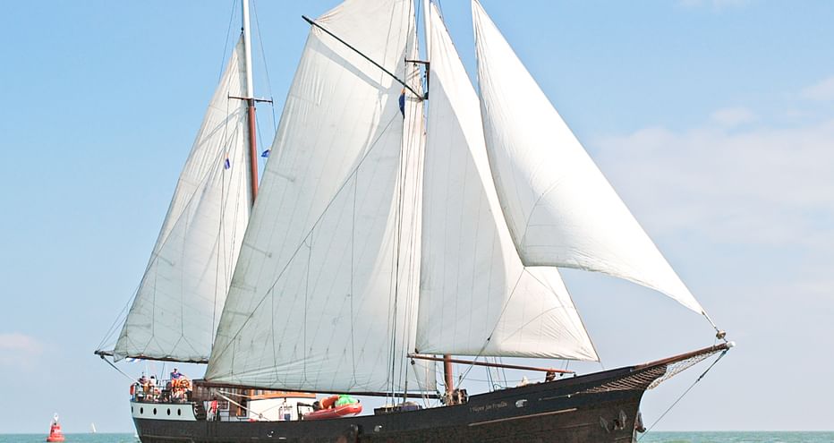 Traditionelles niederländisches Segelschiff mit dunklem Holzrumpf und weißen Segeln auf ruhigem Wasser unter blauem Himmel.