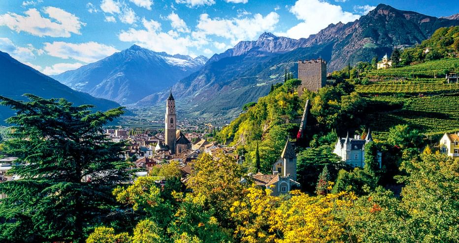 Malerischer Blick auf einen Südtiroler Ort mit Kirchturm, historischen Gebäuden und Weinbergen an Hängen, umgeben von imposanten Alpenbergen.