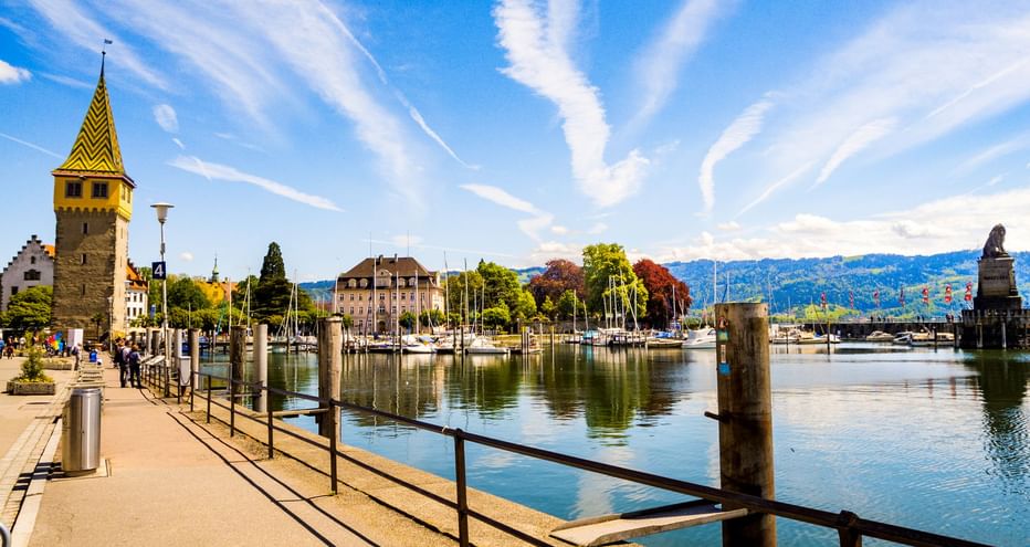 Lindauer Hafenpromenade mit historischem Leuchtturm, vertäuten Segelbooten und Löwenstatue am Hafeneingang unter blauem Himmel mit Bergen im Hintergrund.
