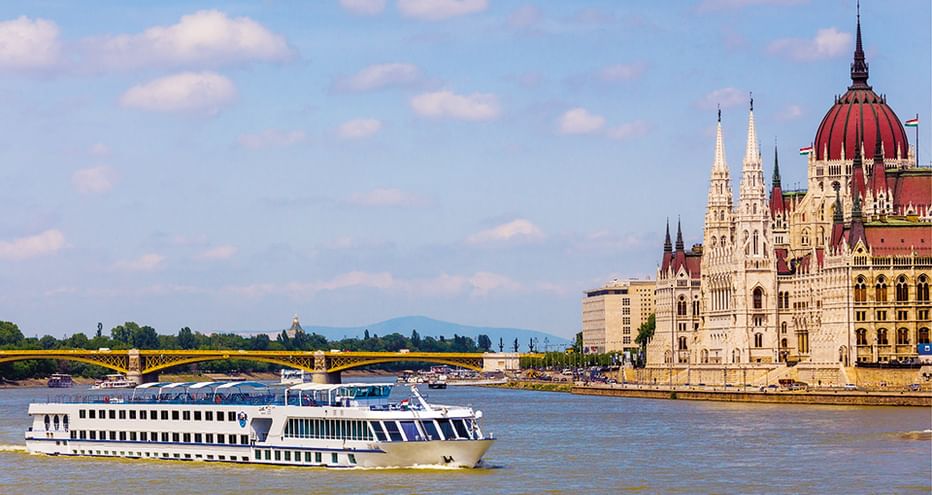 Weißes Flusskreuzfahrtschiff auf der Donau mit dem ungarischen Parlamentsgebäude und roter Kuppel am Flussufer in Budapest.