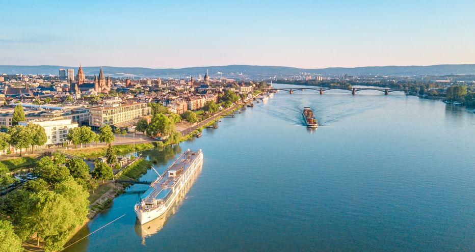 Luftaufnahme eines Flusskreuzfahrtschiffs auf dem Main in Mainz mit Stadtpanorama, Brücken und grünen Ufern unter klarem Himmel.