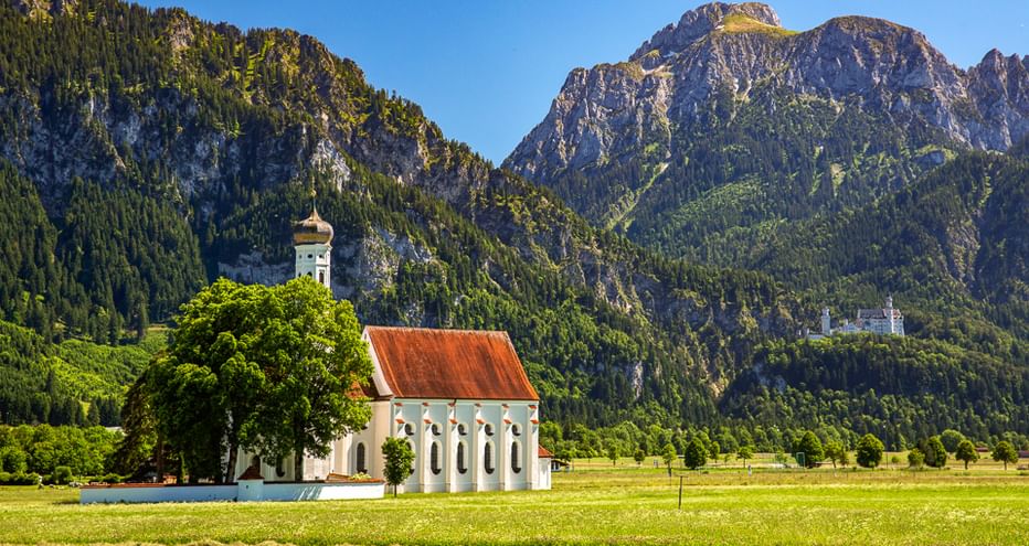 Weiße Kapelle mit rotem Dach auf grüner Wiese, umgeben von bewaldeten Bergen. Schloss Neuschwanstein sichtbar auf entferntem Hang unter blauem Himmel.