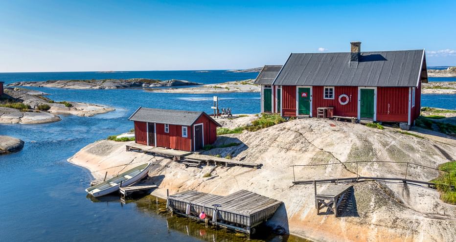 Rote Holzhütten mit grünen Türen an felsiger Küste im Stockholmer Schärengarten. Kleine Boote am Steg vertäut, ruhiges blaues Wasser und Inseln sichtbar.