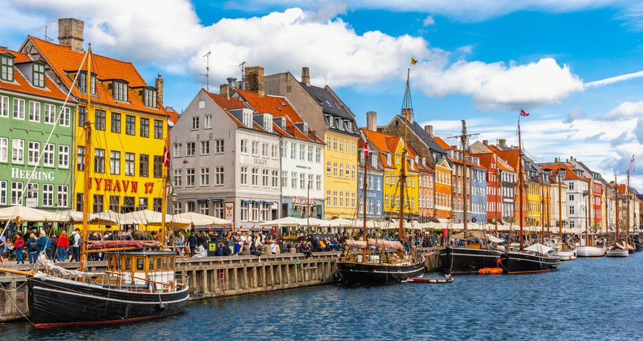 Historische Nyhavn-Uferpromenade in Kopenhagen mit bunten Stadthäusern, traditionellen Holzbooten im Kanal und Besuchern unter blauem Himmel.