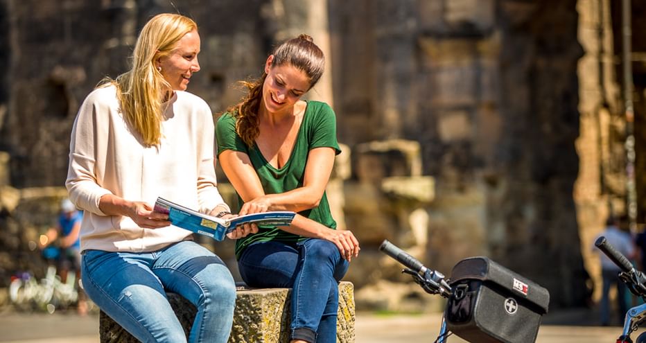 Zwei Frauen sitzen auf einer Steinmauer und studieren eine Karte, Fahrräder neben ihnen. Historisches Steingebäude im Hintergrund.