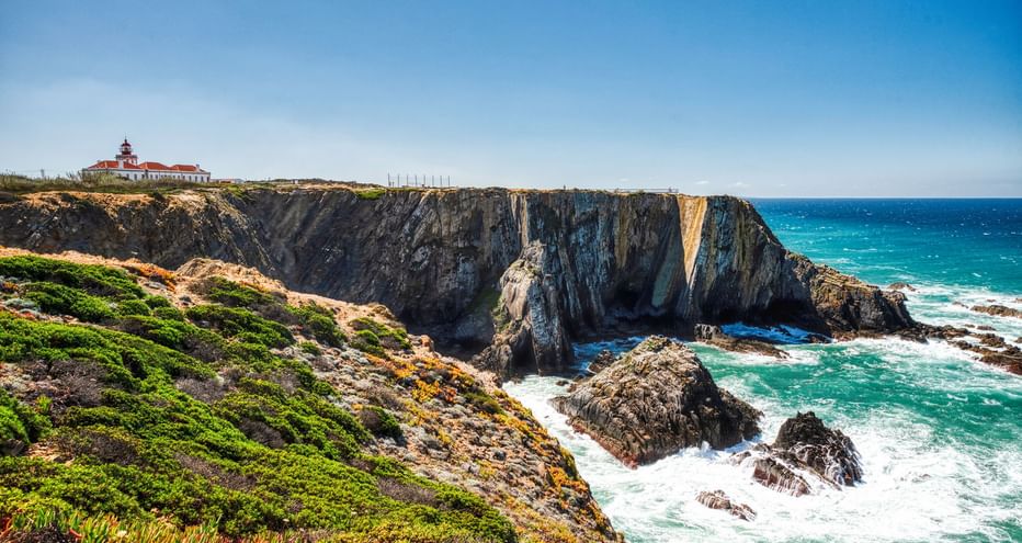 Leuchtturm mit rotem Dach auf dramatischen Felsenklippen an Portugals Atlantikküste. Türkisfarbene Wellen brechen an dunklen Felsen unter grüner Vegetation.