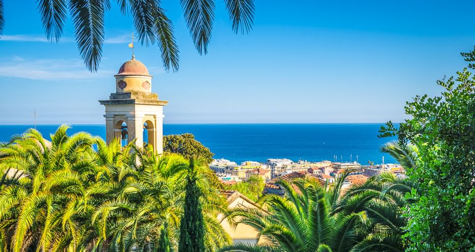 Weißer Glockenturm mit Kuppel, umrahmt von Palmen, mit Blick auf eine Küstenstadt an der italienischen Riviera und blauem Mittelmeer.