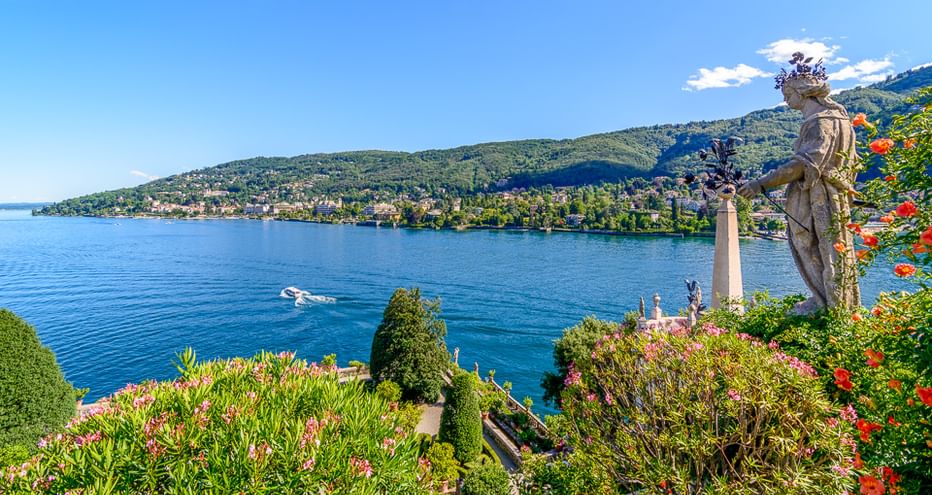 Klassische Statue in einem blühenden Garten mit Blick auf den Lago Maggiore mit grünen Hügeln und blauem Wasser. Ein Boot ist auf dem See sichtbar.