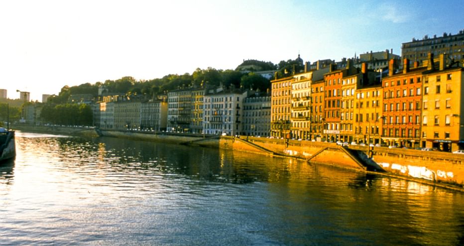 Blick auf die Rhône in Lyon mit farbenfrohen historischen Gebäuden entlang des Ufers, die von goldenem Sonnenlicht beleuchtet werden.