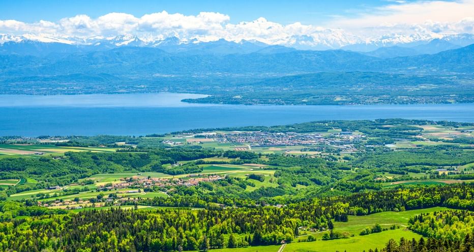Panoramablick vom Jura Vaudois mit grünen Wäldern und Feldern, einem großen blauen See und schneebedeckten Alpen unter blauem Himmel mit Wolken.