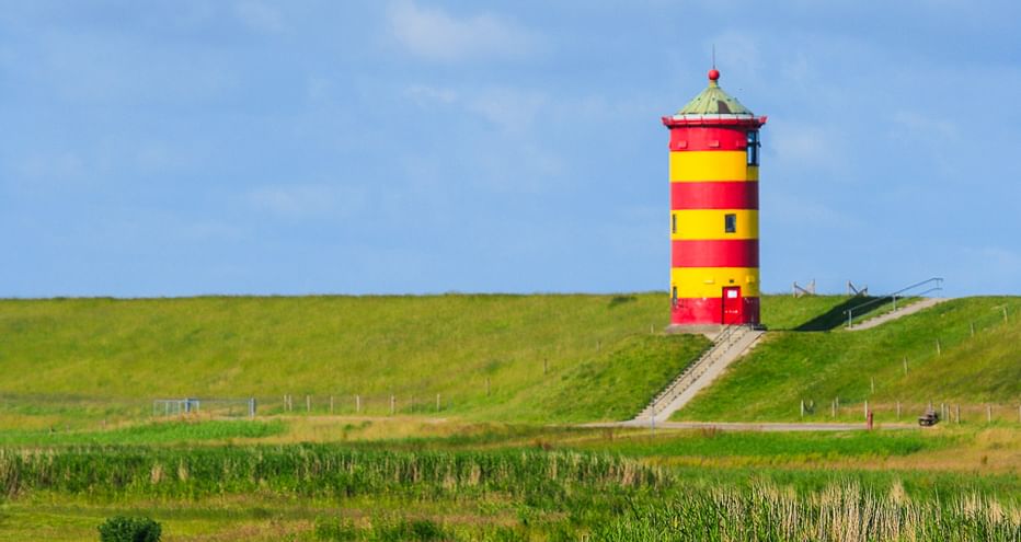 Rot-gelb gestreifter Leuchtturm auf grünem Deich mit Weg. Marschland im Vordergrund, blauer Himmel darüber.