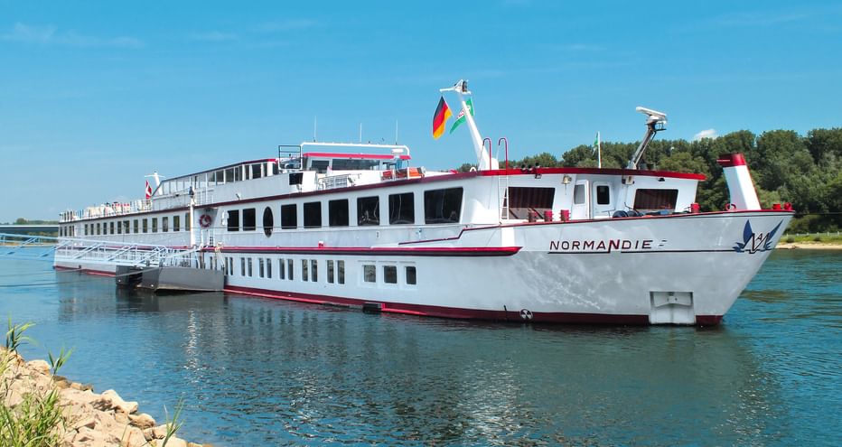 Weißes Flusskreuzfahrtschiff Normandie mit rotem Zierstreifen auf blauem Wasser. Das mehrstöckige Schiff zeigt eine deutsche Flagge vor klarem Himmel.