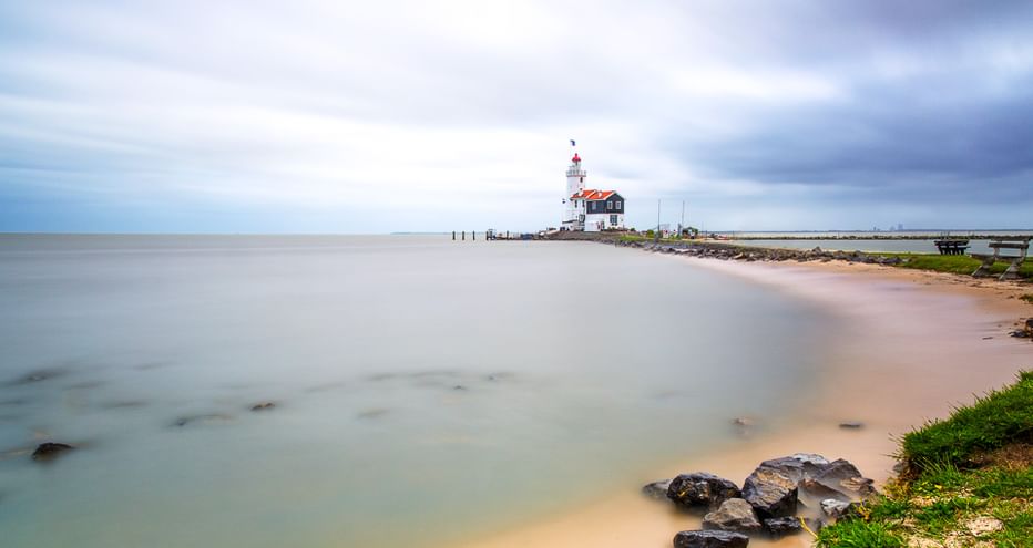 Weißer Leuchtturm mit rotem Dach auf einem Steg am IJsselmeer. Sandstrand mit Steinen im Vordergrund unter bewölktem Himmel.