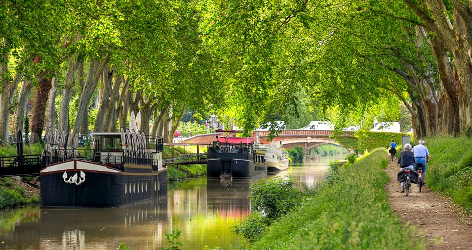 Zwei Radfahrer auf einem von Bäumen gesäumten Treidelpfad am Canal du Midi mit vertäuten Booten und einer Brücke im Hintergrund.