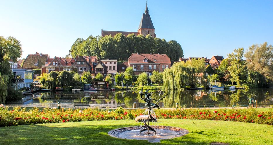 Blick über den Mölln Stadtsee mit Brunnen im Vordergrund, bunten Blumenbeeten, historischen Häusern am Ufer und Kirche auf dem Hügel.
