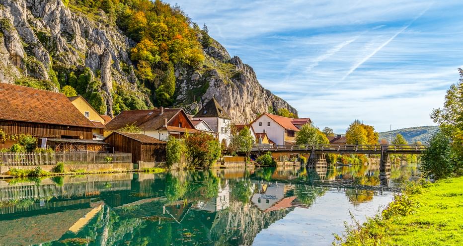 Essing village along the Altmühl river with traditional houses, wooden footbridge, and dramatic limestone cliffs in autumn colors.
