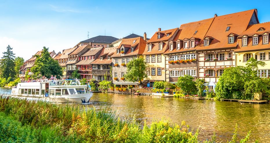 Colorful half-timbered houses with orange roofs along the Main River. A tour boat is moored at the waterfront, with green vegetation in foreground.