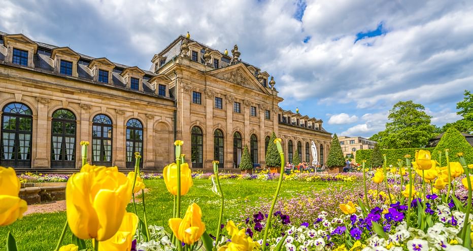 Baroque palace with ornate facade and blooming garden featuring yellow tulips and purple flowers under a cloudy blue sky.