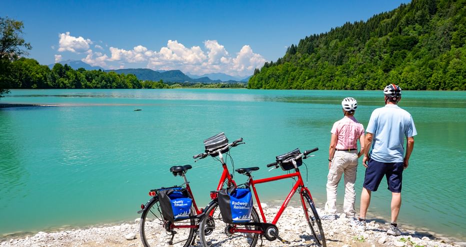 Zwei Radfahrer stehen am Ufer des türkisfarbenen Feistritzer Stausees mit zwei roten Tourenrädern, bewaldete Hügel und Berge im Hintergrund.